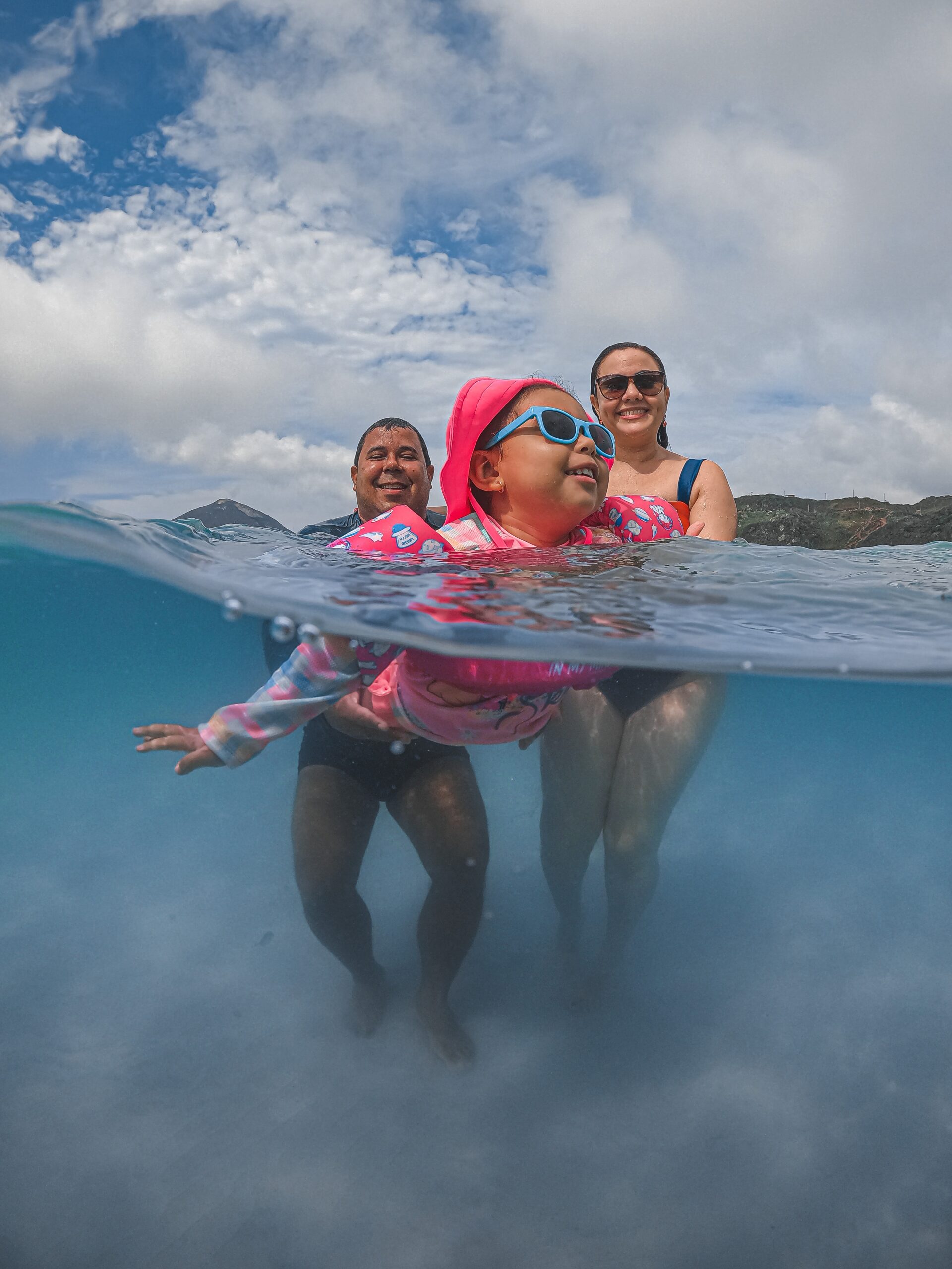 Young girl swims with her parents behind her. Camera is half in the water.