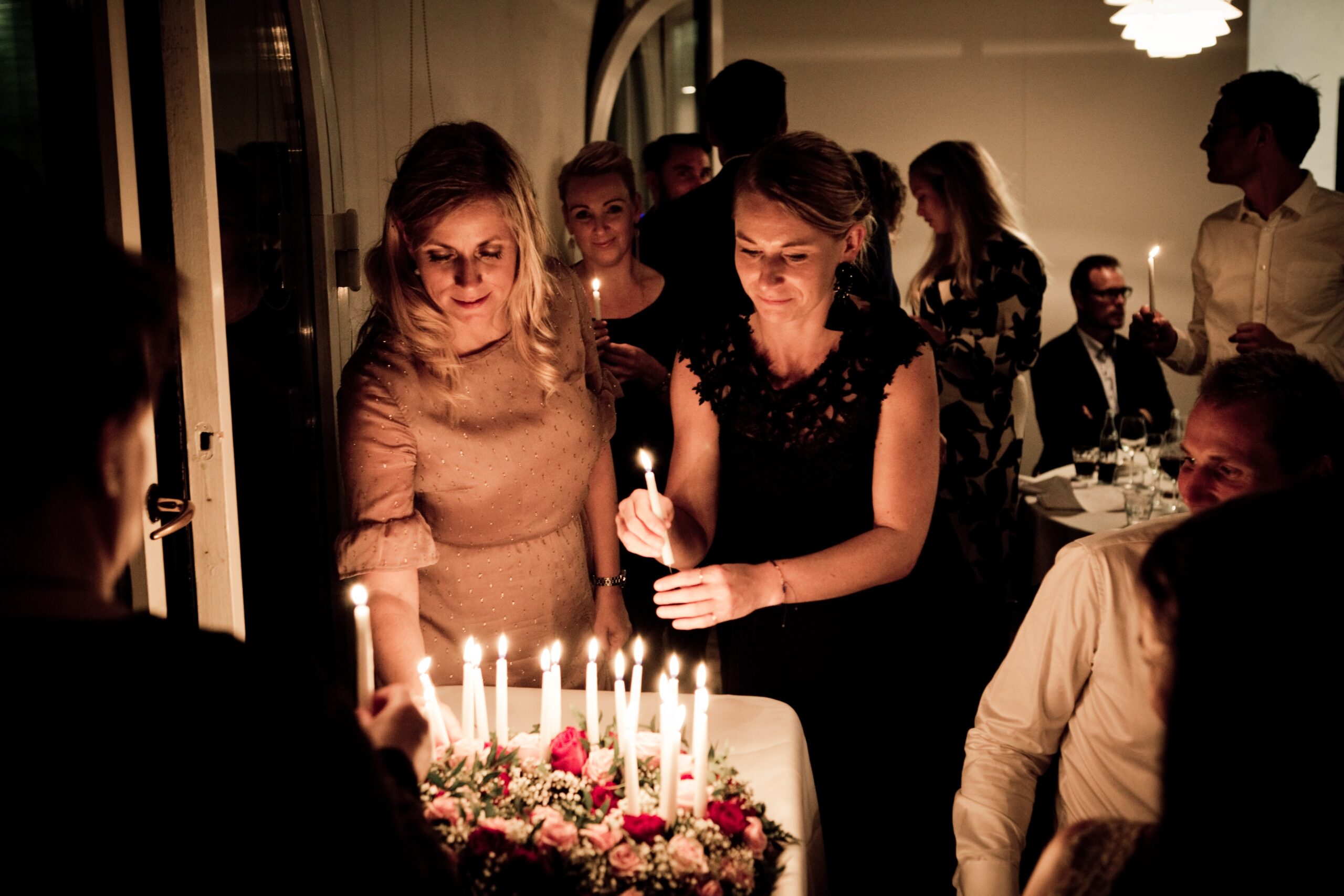Two women light a birthday cake at a dimly lit celebration.