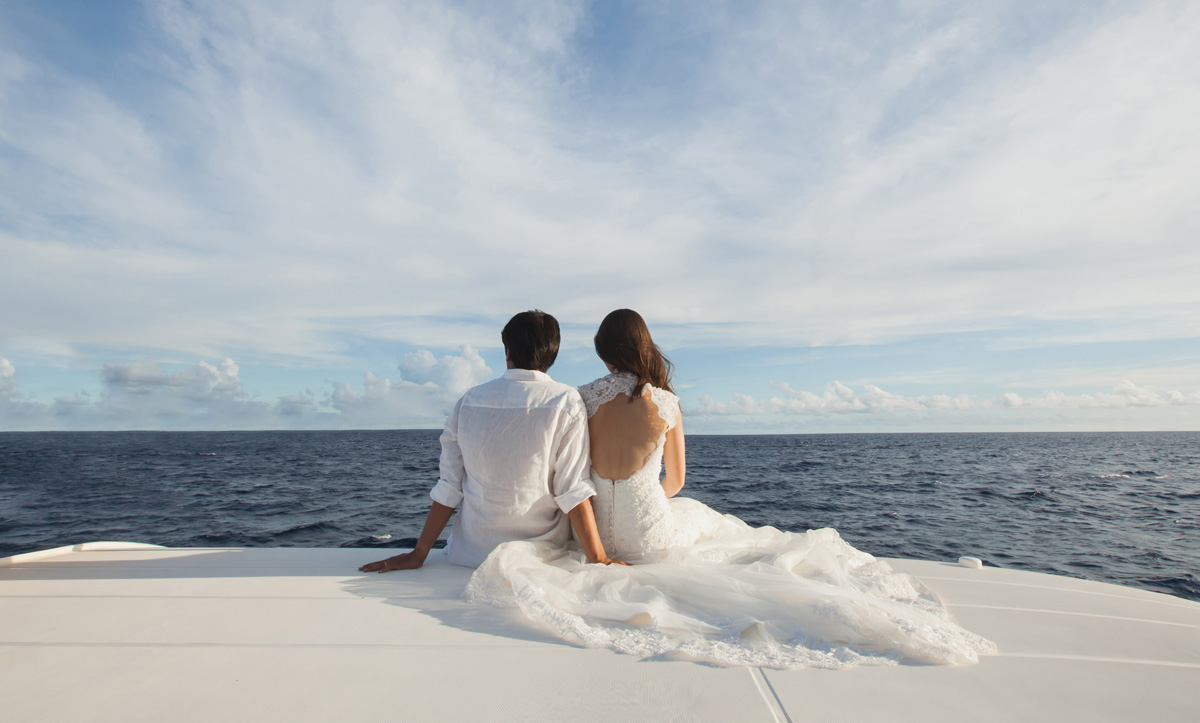 Bridge and groom sit on the edge of a yacht looking out at the ocean.